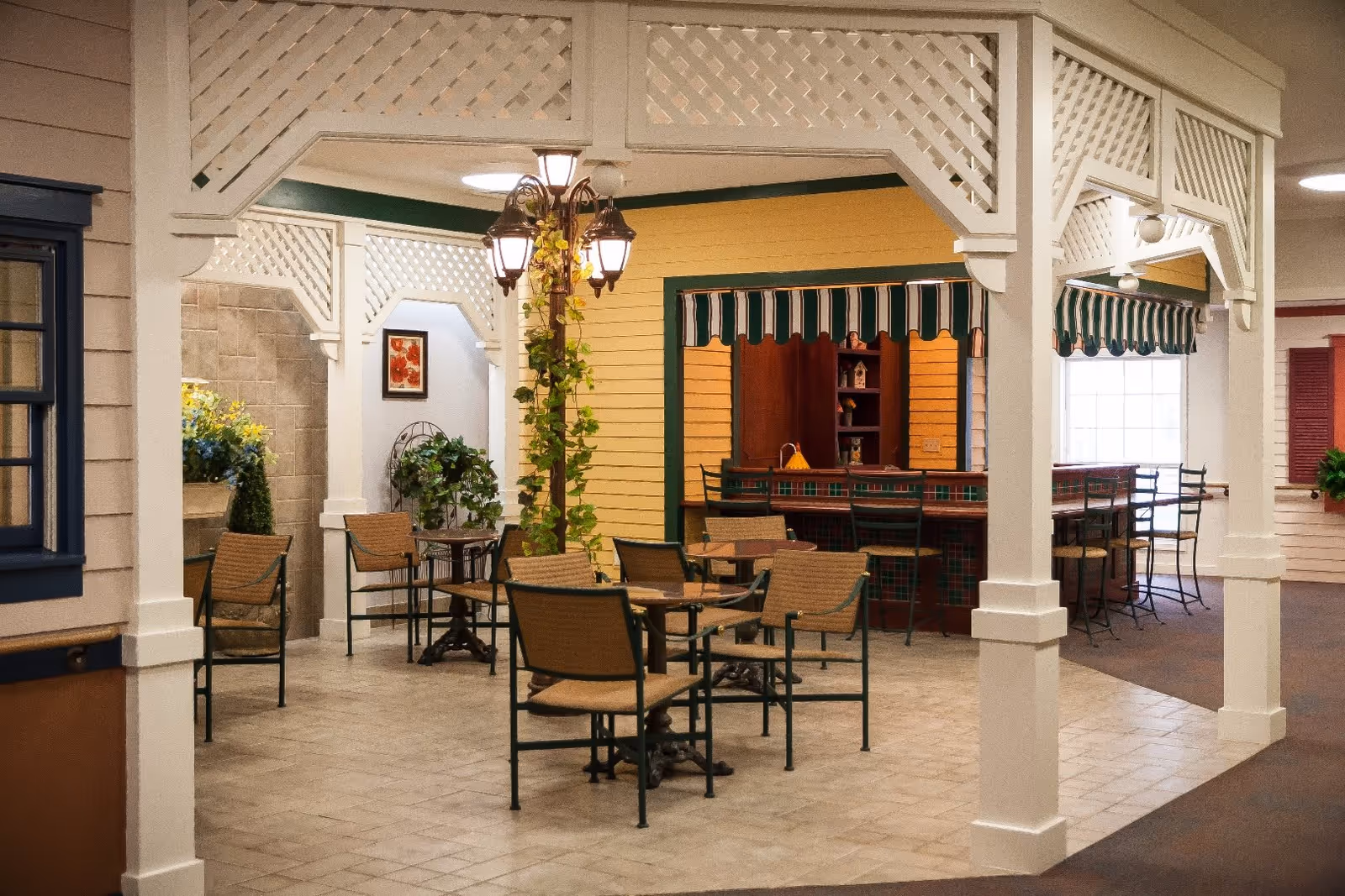 Indoor seating area designed to resemble an outdoor patio with several tables and chairs, a lamppost with hanging plants, and a bar counter with high stools under a striped awning. The space features white lattice columns and decorative elements, with potted plants and framed artwork on the walls.