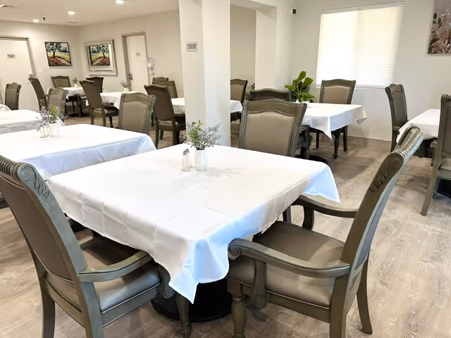 A dining room with multiple tables covered in white tablecloths, each surrounded by cushioned wooden chairs. Small floral centerpieces and salt and pepper shakers are placed on the tables. The room has light-colored walls, framed artwork, and a window with a blind partially drawn.