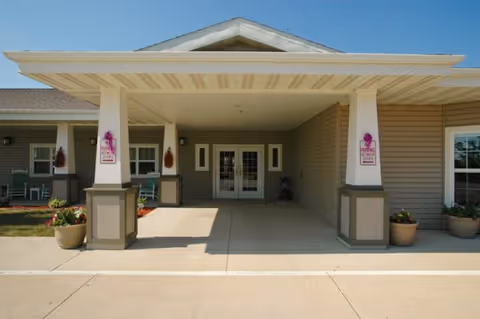 Entrance of a senior living facility with a covered drop-off area supported by columns. The building exterior is beige with white trim, and there are potted plants on either side of the entrance. Two signs with pink ribbons are mounted on the columns, indicating a breast cancer awareness theme.