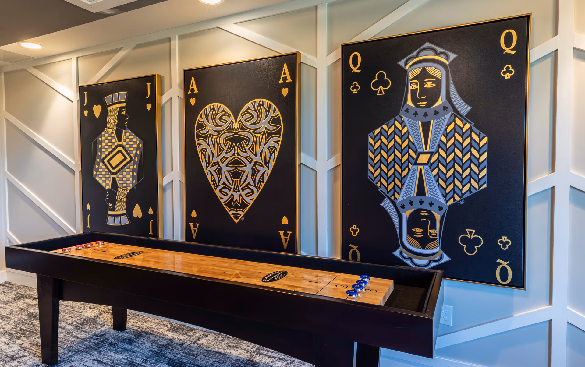 A shuffleboard table with red and blue pucks in a room decorated with three large playing card artworks on the wall, featuring the Jack of Hearts, Ace of Hearts, and Queen of Clubs in black, gold, and blue colors.