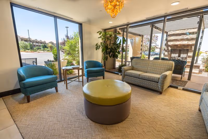 Sunny seating area with teal armchairs, a patterned sofa, and a round green ottoman in front of large windows and a patio.