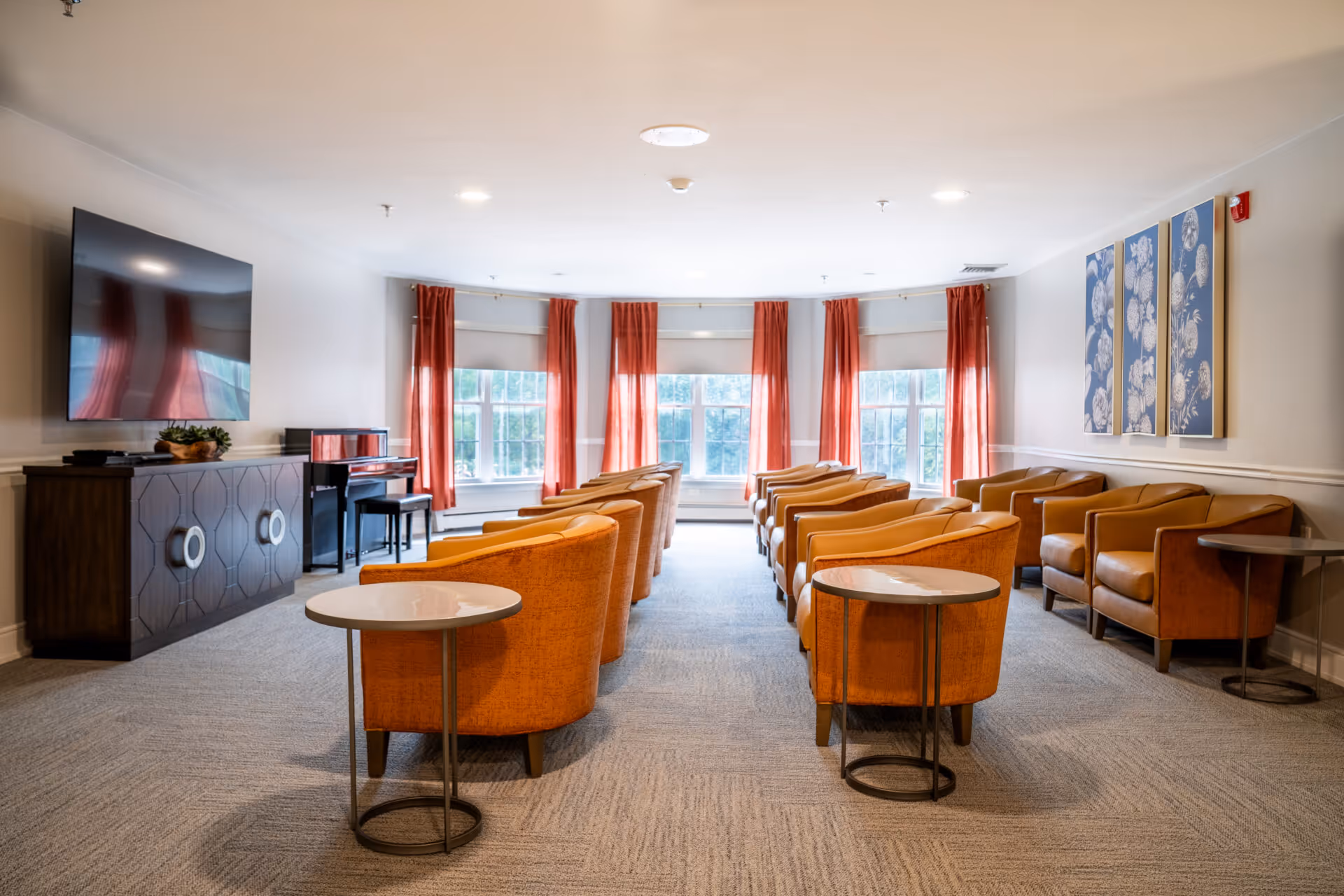 A bright and spacious living room with two rows of orange armchairs facing a large flat-screen TV mounted on the wall. The room has large windows with red curtains allowing natural light to fill the space. There is a dark wooden cabinet under the TV and a piano with a bench near the windows. Three framed floral artworks hang on the right wall. Small round side tables are placed beside some of the chairs.