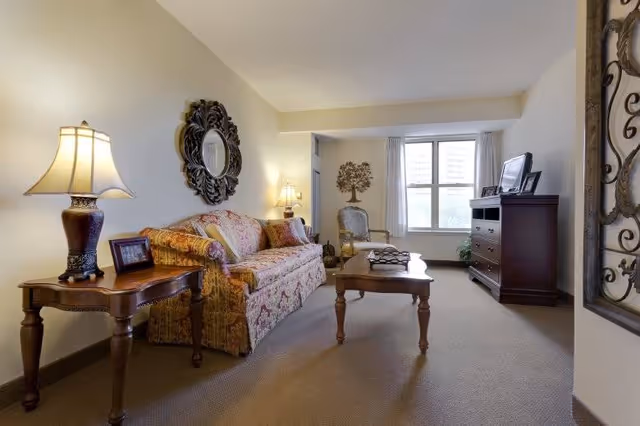 A cozy living room in a senior living facility featuring a patterned sofa with cushions, a wooden coffee table, a side table with a lamp and framed photo, an armchair near a window with white curtains, a wooden dresser with a TV and picture frames on top, and decorative wall art including a round mirror and a tree sculpture.