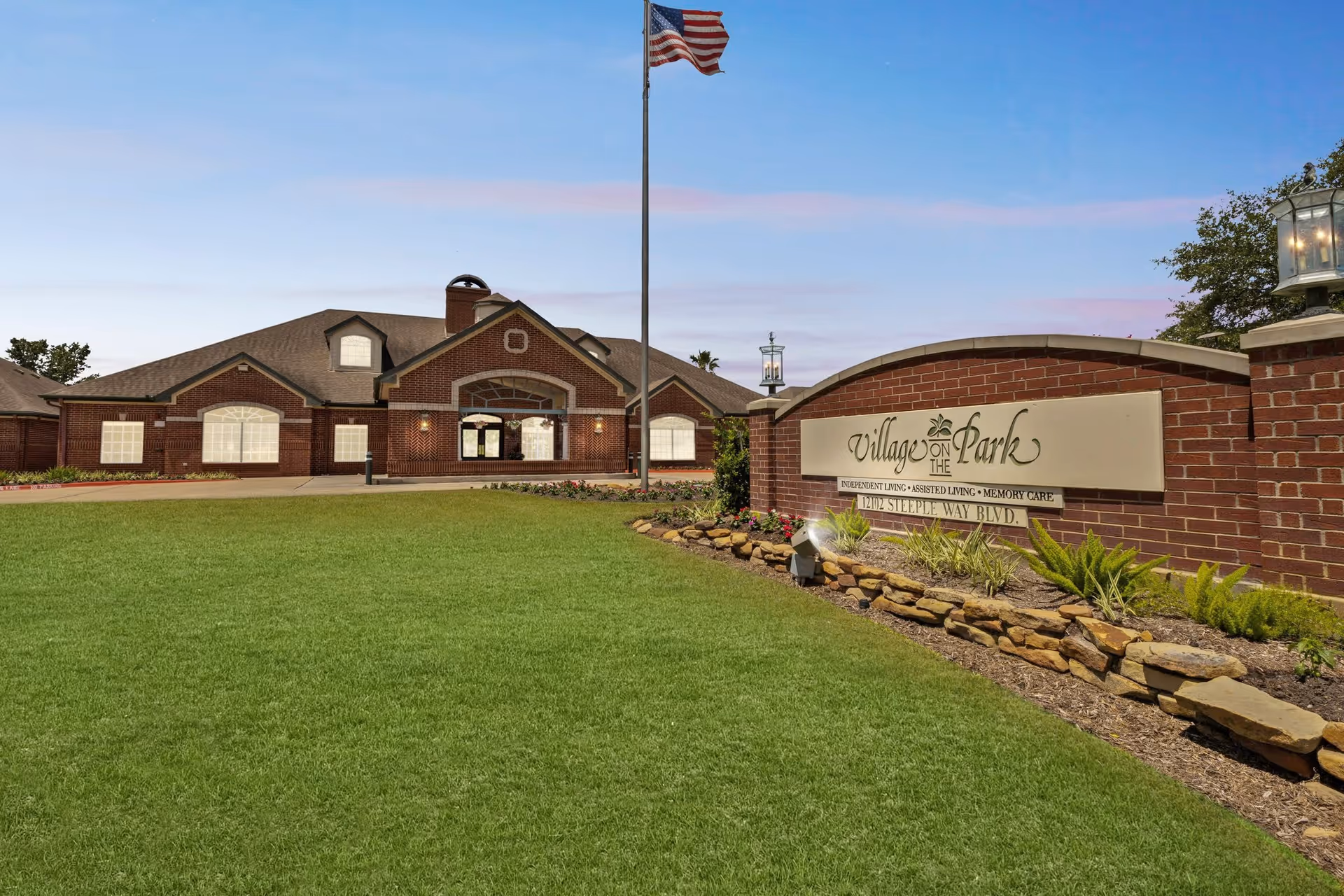 Exterior view of Village on the Park Steeplechase senior living facility with a large green lawn, a brick building with multiple windows, and an American flag on a flagpole. A brick sign in the foreground displays the facility name and services offered.