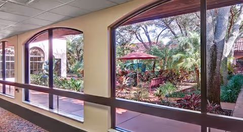 Interior corridor with large arched windows overlooking a landscaped courtyard with trees, plants, and a red umbrella.
