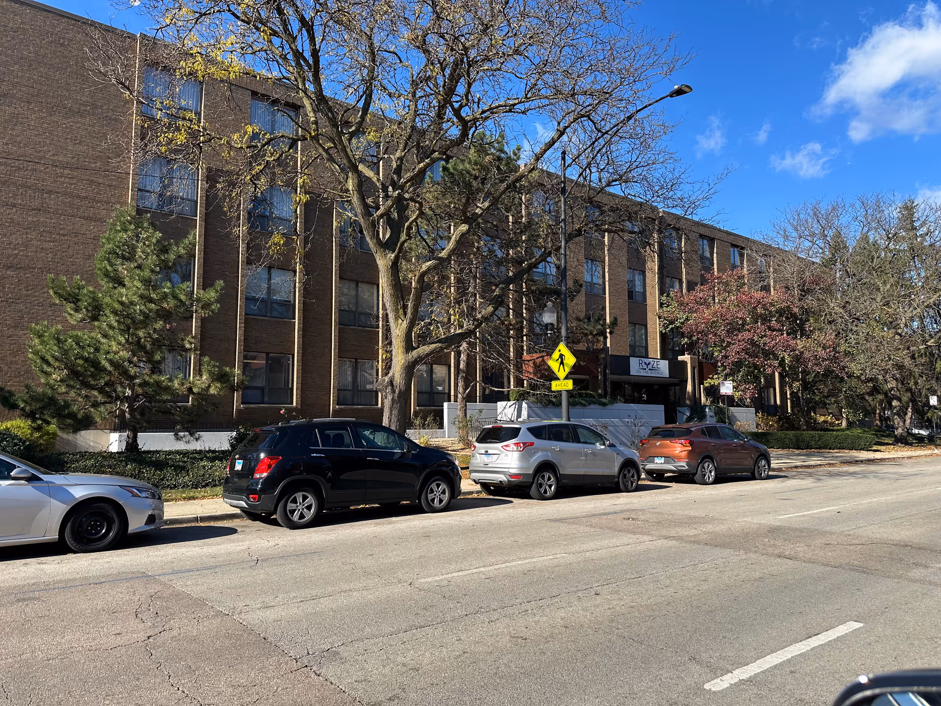 Four-story brick building front with a 'RYZE' entrance, trees, and several cars parked along the street under a blue sky.