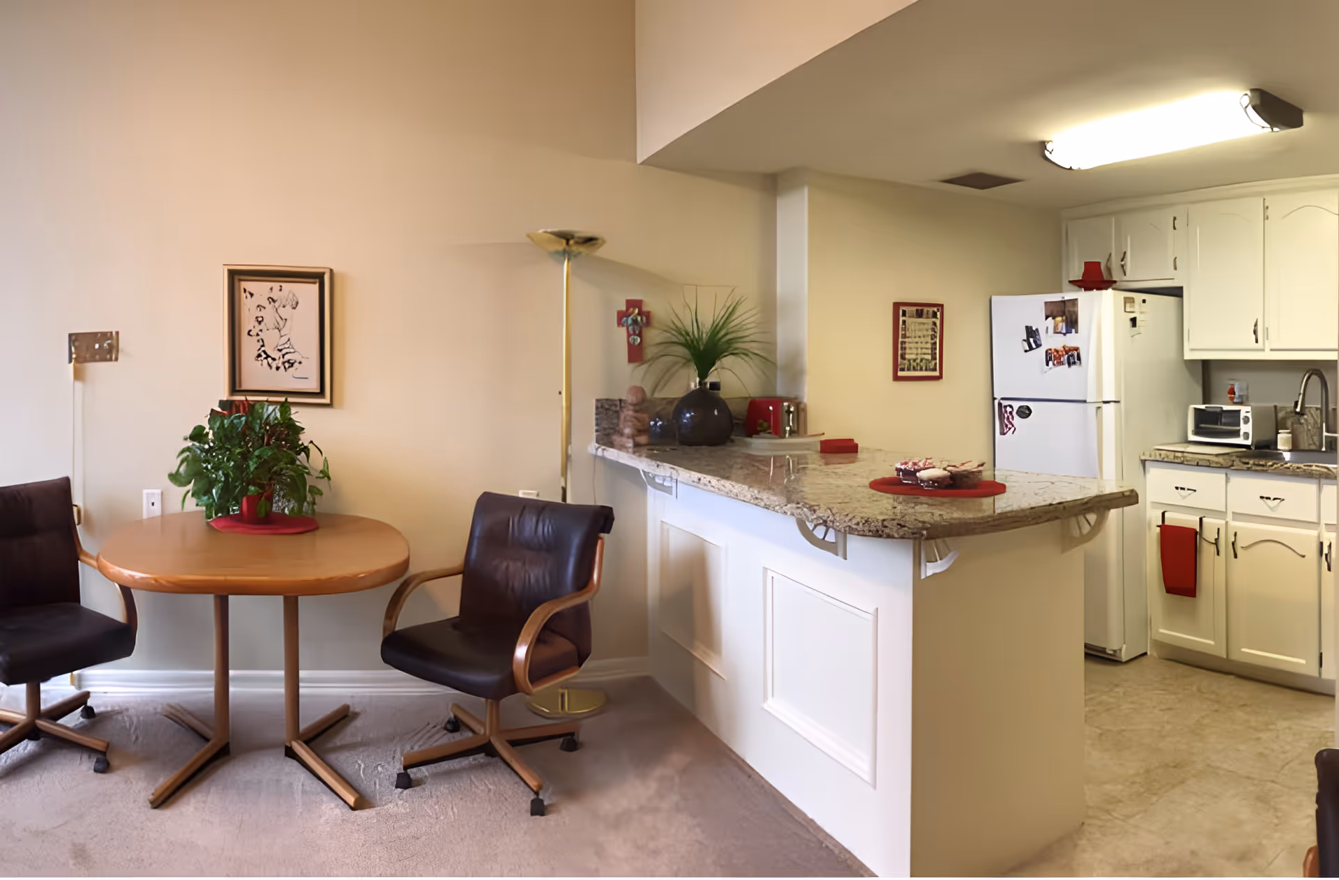 A cozy kitchen and dining area in a senior living facility. The dining area features a small round wooden table with two black cushioned chairs on wheels. A green potted plant sits on the table. The kitchen has white cabinets, a granite countertop with decorative items, a white refrigerator with magnets and photos, a microwave, and a sink. The walls are painted beige, and there is a floor lamp next to the dining table.