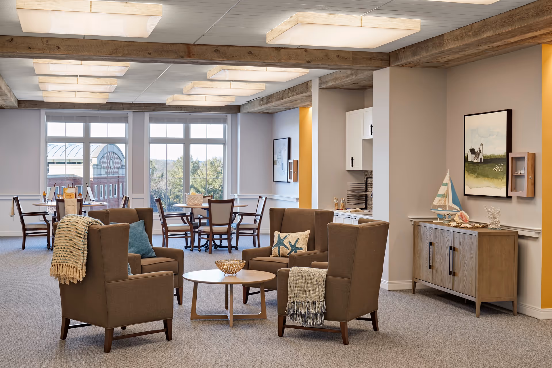 A bright and spacious common area in a senior living facility featuring four brown armchairs arranged around a round wooden coffee table. The chairs have decorative pillows and throws. Behind the seating area, there are two round tables with chairs near large windows that let in natural light and offer a view of trees and a building outside. The room has exposed wooden beams on the ceiling, modern rectangular light fixtures, and a sideboard with nautical decor and a framed painting on the wall.