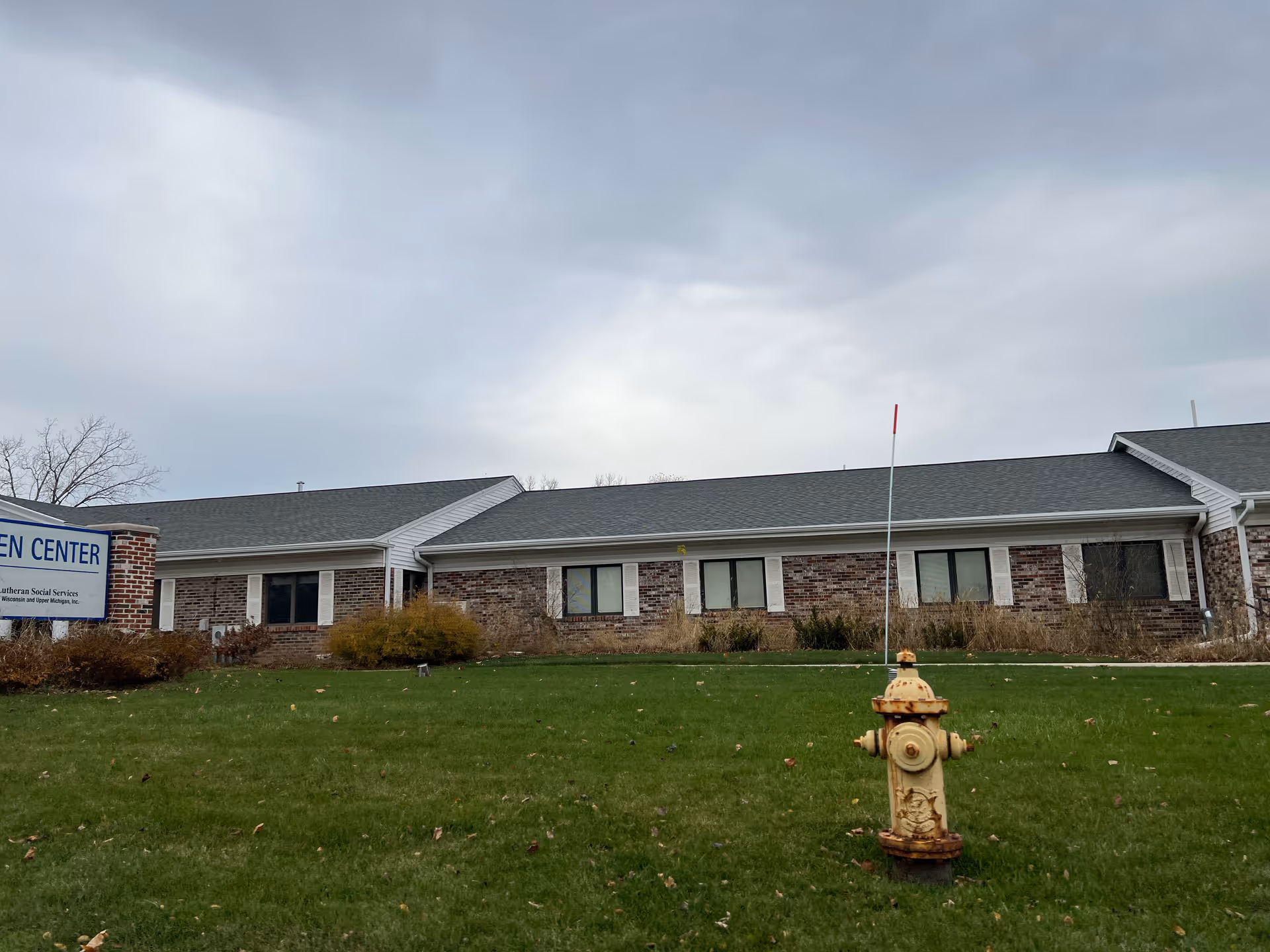 Exterior view of a single-story brick building with white window shutters and a gray roof under a cloudy sky. In front of the building is a green lawn with a yellow fire hydrant and a sign partially visible on the left side that reads 'CENTER'.