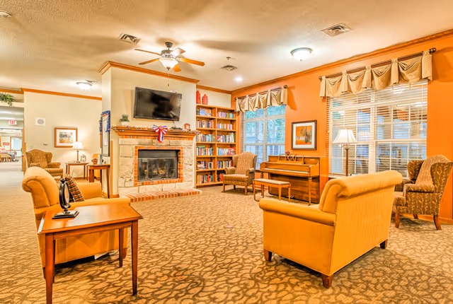 Cozy sitting area with upholstered sofas and armchairs around a fireplace topped by a wall-mounted TV, bookshelves, a piano and large windows.
