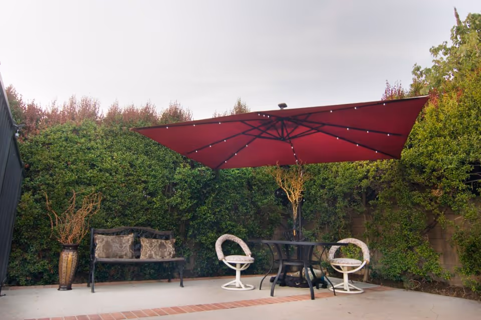 Outdoor patio area with a large red umbrella, two cushioned chairs, a black metal table, a bench with two decorative pillows, and a tall vase with dried branches. The area is surrounded by green bushes and a brick border on the ground.