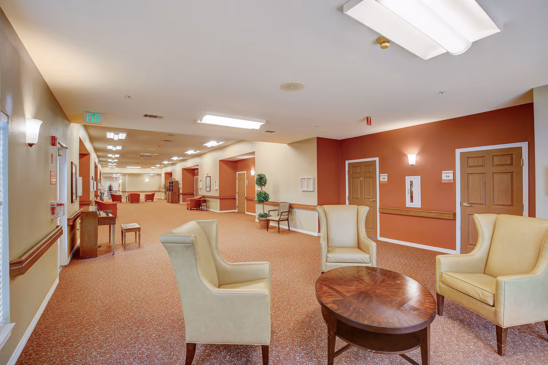 A spacious and well-lit hallway in a senior living facility with beige and rust-colored walls. The carpet is patterned in shades of brown and orange. There are three beige armchairs arranged around a wooden oval coffee table in the foreground. Along the hallway, there are wooden doors with room numbers, wall-mounted lights, a piano with a small bench, and some chairs and plants placed intermittently. The ceiling has multiple rectangular fluorescent light fixtures and an exit sign is visible on the left side.