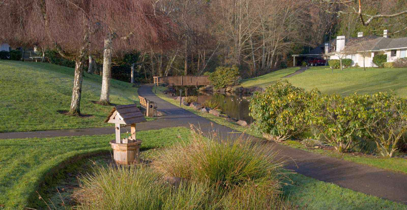Landscaped outdoor path winding past a small decorative wishing well, pond and wooden footbridge with buildings and trees in the background.
