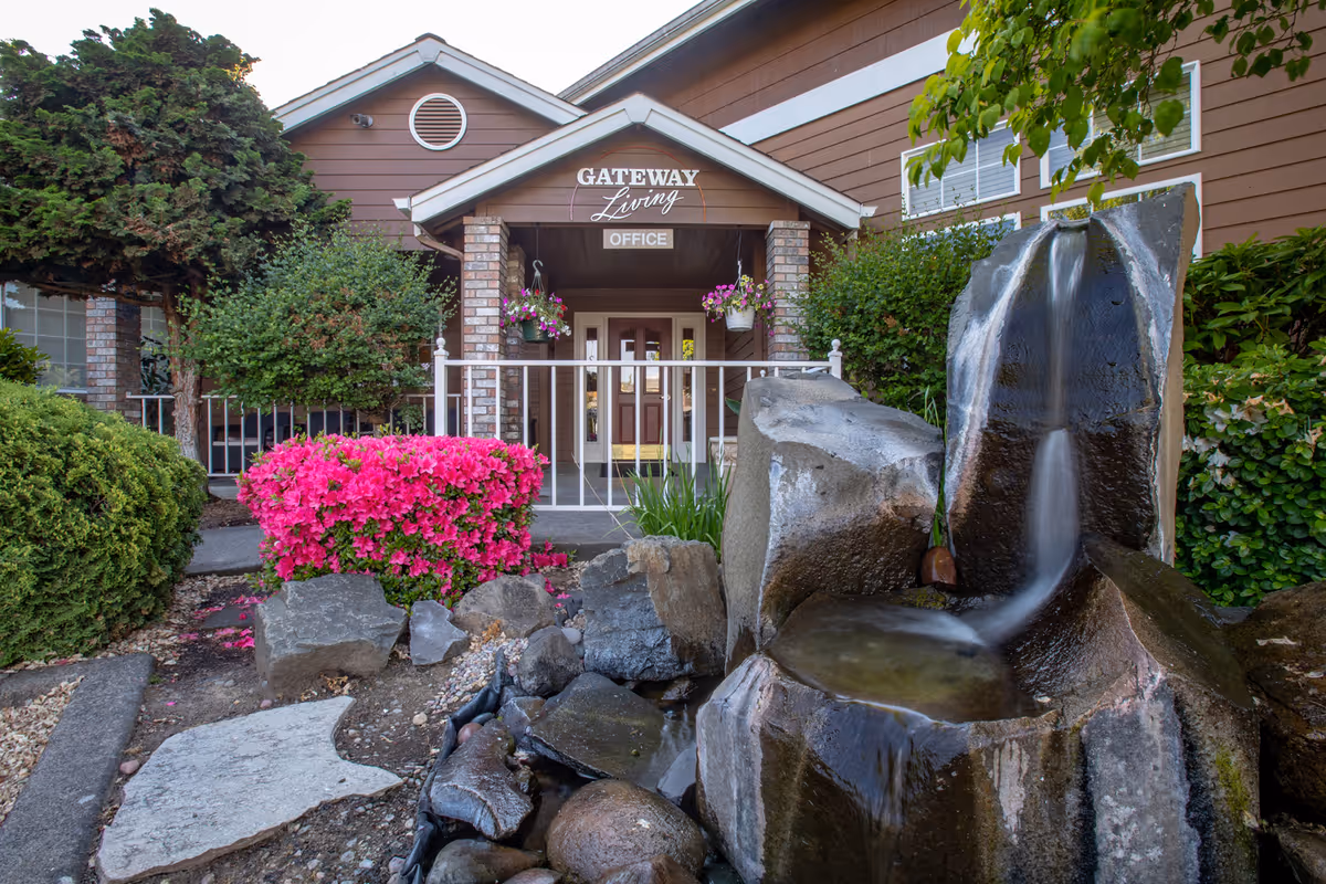 Entrance to Gateway Living office with a brown building, white railing, hanging flower pots, and a water fountain surrounded by rocks and greenery including bright pink flowers.