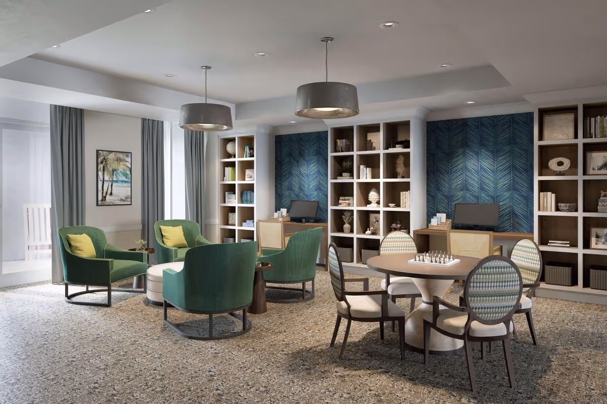 A cozy common area in a senior living facility featuring a round table with four chairs and a chessboard on top. There are four green armchairs arranged around a small round ottoman. The room has built-in bookshelves with decorative items and books, two computer desks, and large windows with curtains. Two modern pendant lights hang from the ceiling.