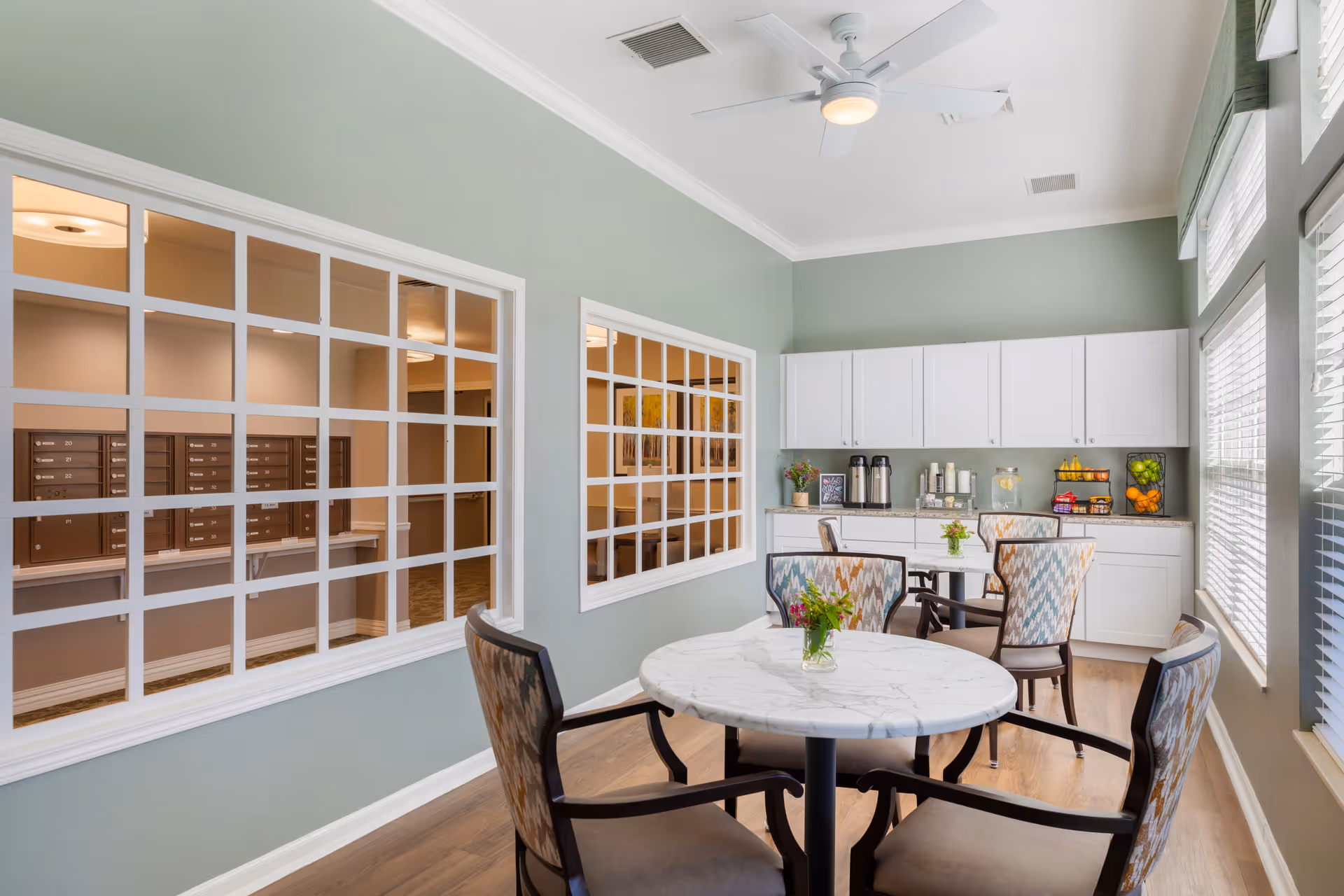A bright and cozy common area with round marble-top tables and patterned chairs. The room has light green walls, large windows with blinds, white cabinets, and a countertop with coffee dispensers, water with lemon, and fruit baskets. Two large windows with white grid frames look into an adjacent room with mailboxes.
