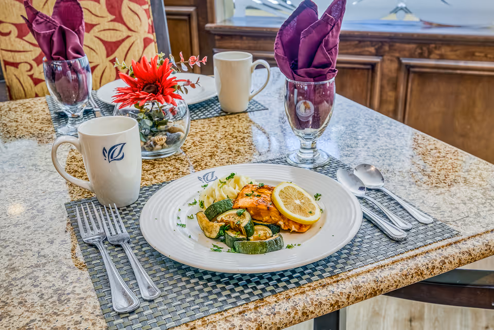 A dining table set with a plate of cooked salmon topped with a lemon slice, mashed potatoes, and sautéed zucchini. The table also has two white mugs, two glasses with folded purple napkins, silverware, and a small glass vase with a red flower and decorative stones. The table surface is a speckled granite, and there is a cushioned chair with a red and gold patterned backrest.