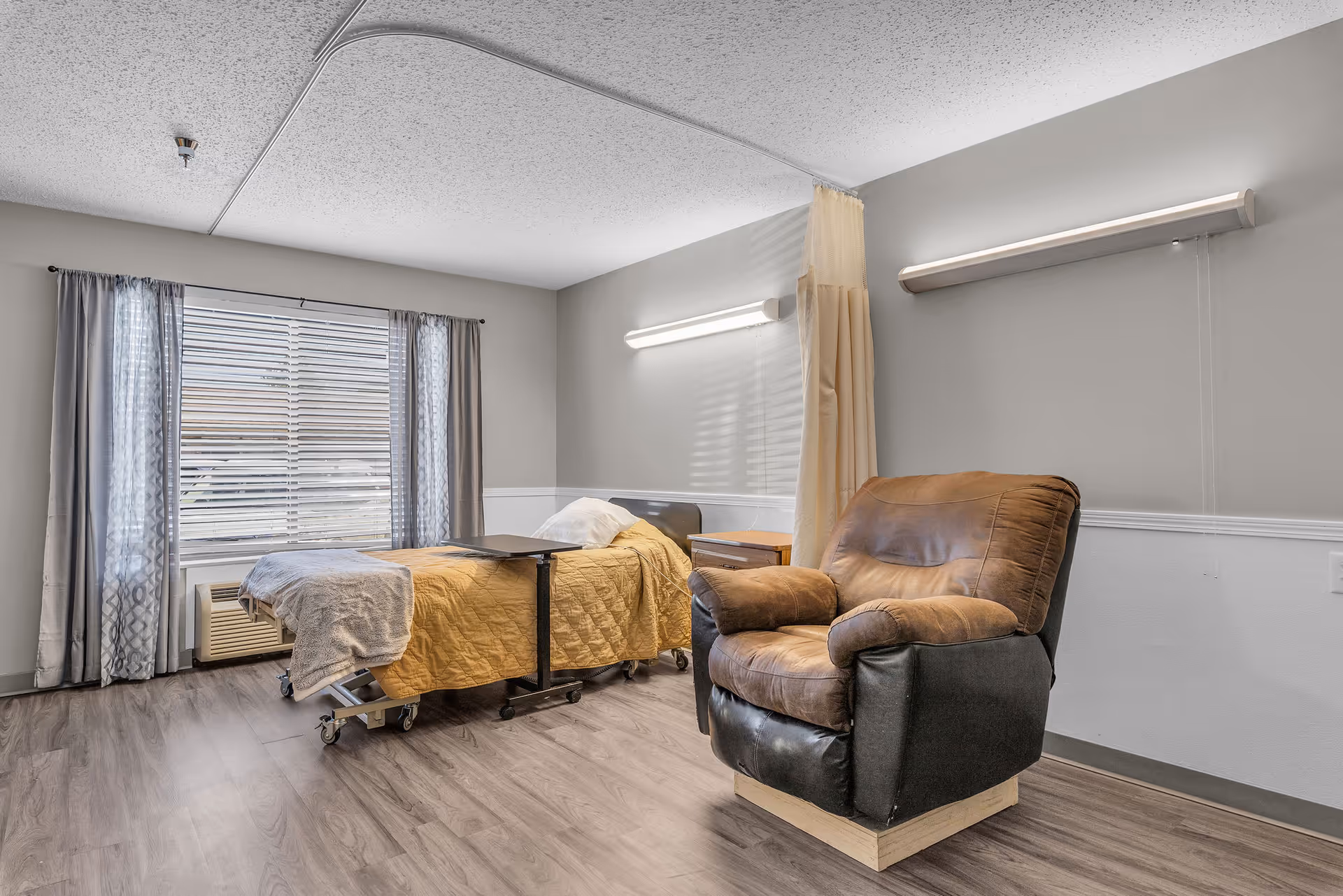 A senior-care bedroom featuring a medical bed with an overbed table, a leather recliner, window blinds with curtains, and a privacy curtain.