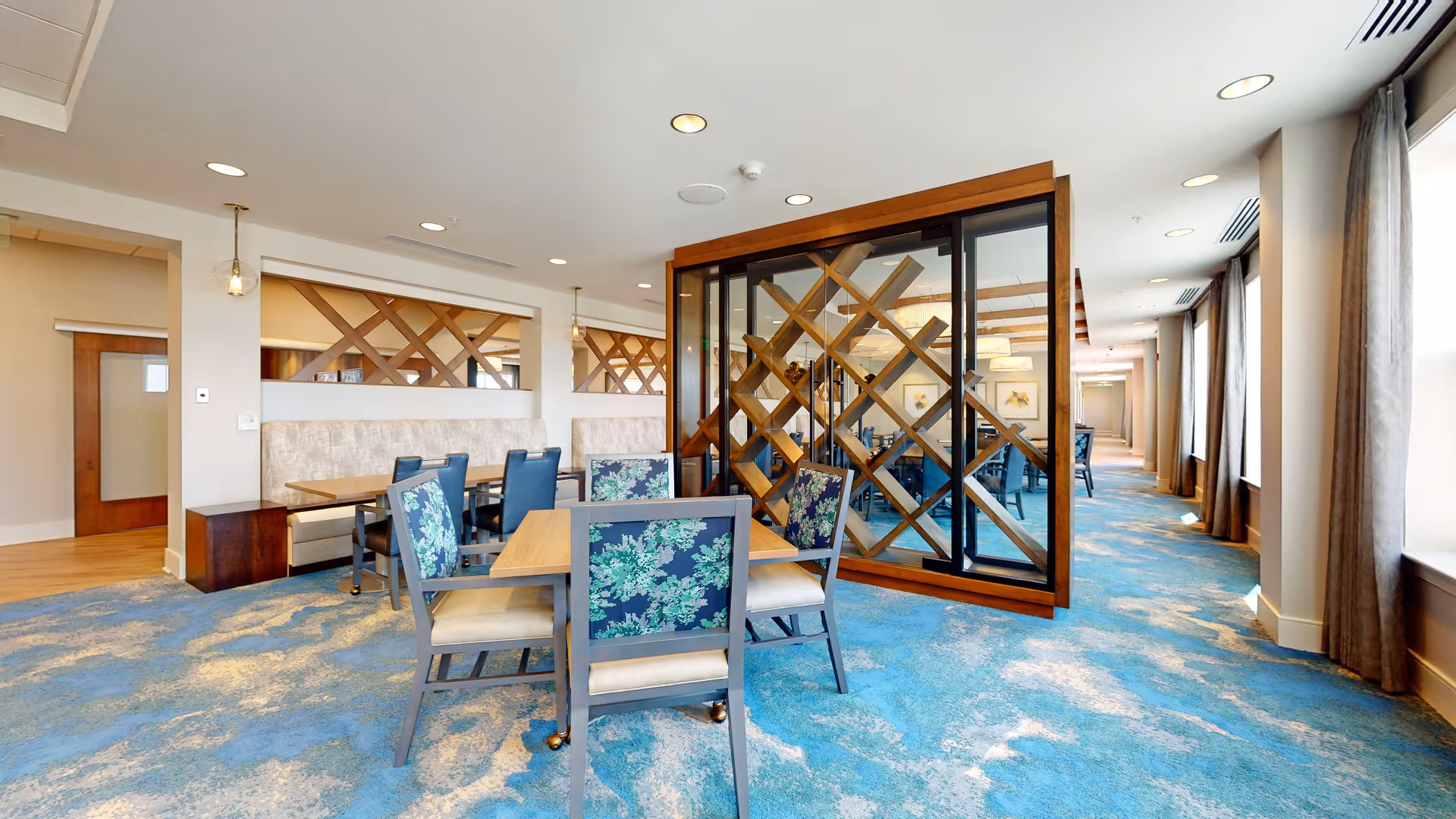 A bright dining area with blue and beige patterned carpet, wooden tables, and chairs with floral upholstery. There is a glass partition with a wooden lattice design separating the space. Large windows with curtains line the right side, allowing natural light to fill the room.