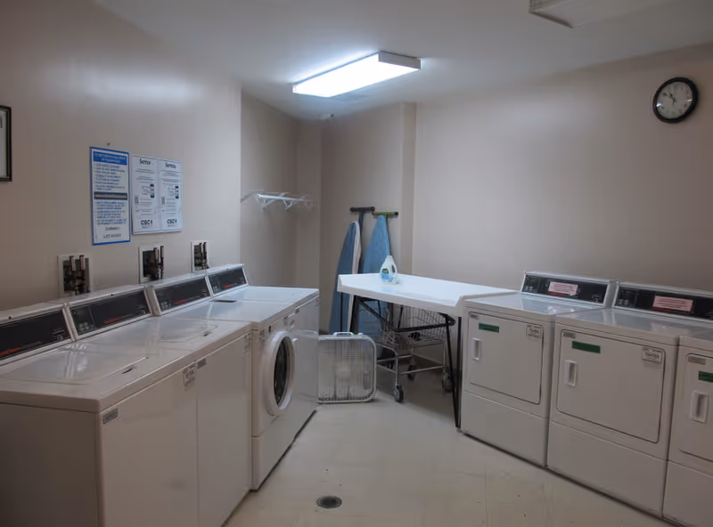 Laundry room with multiple washing machines and dryers along the walls, a folding table with laundry detergent on it, two ironing boards, and a box fan on the floor. The room has beige walls, a clock on the wall, and fluorescent ceiling lighting.