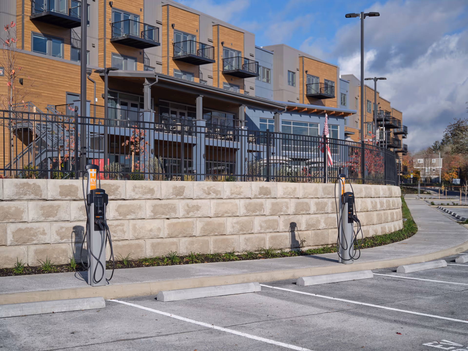 Parking lot with electric vehicle charging stations in front of a multi-story building with balconies and a fenced patio area under a partly cloudy sky.