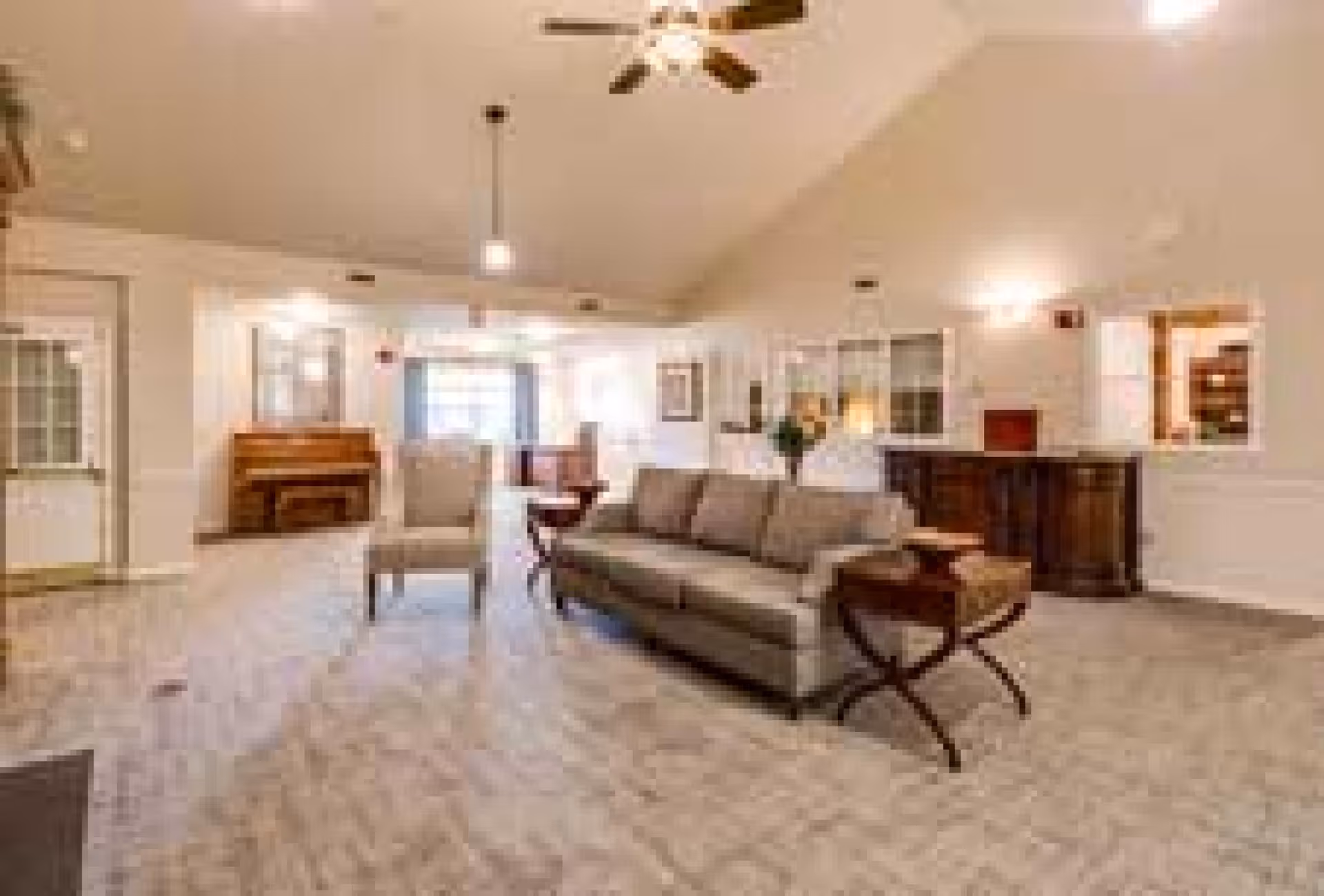 A spacious living room area with a beige sofa and armchair, a wooden side table, a ceiling fan with lights, a piano against the far wall, and large windows allowing natural light to fill the room. The room has light-colored walls and carpeted flooring.