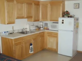 A small kitchen area with light wood cabinets, a white countertop, a sink, a microwave, and a white refrigerator. There are various kitchen items on the counter including a coffee maker and a toaster. A towel hangs from one of the cabinet handles.
