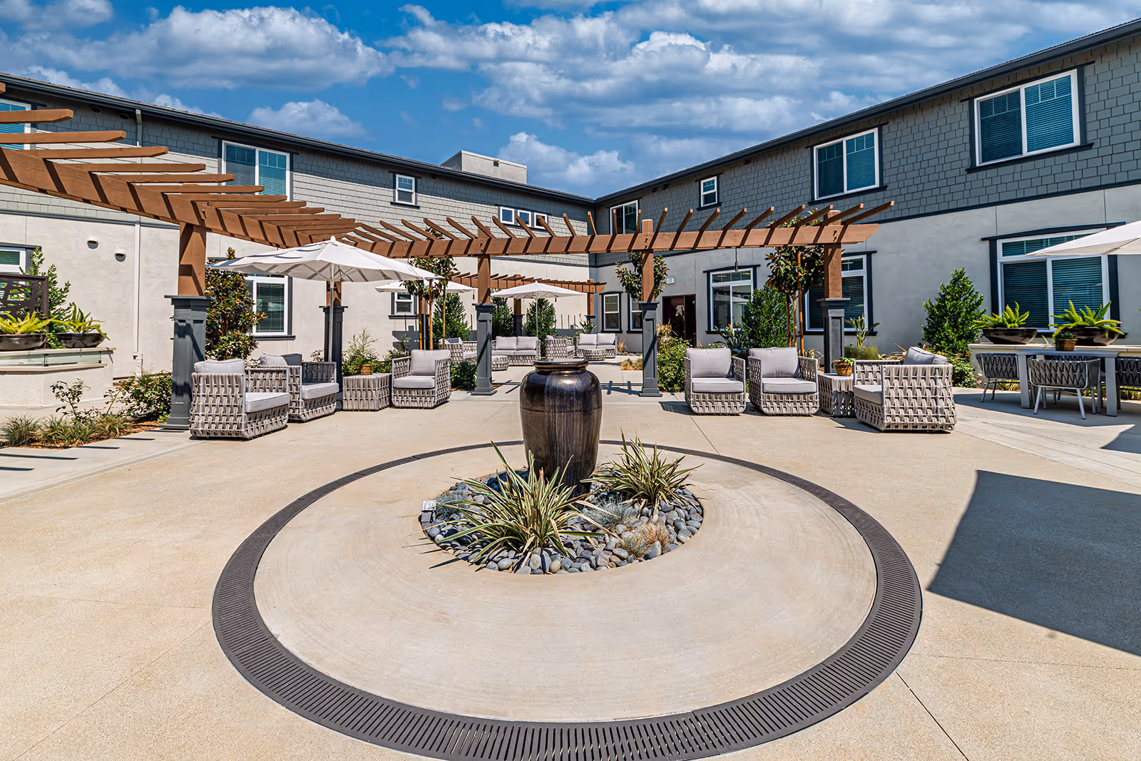 Sunny outdoor courtyard with a circular central planter and fountain, patio seating, pergolas, and umbrellas in front of a two-story senior living building.