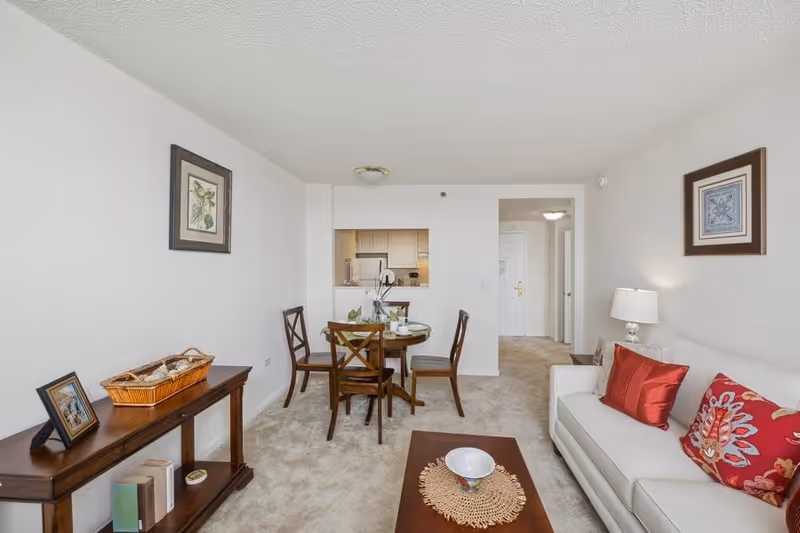 A cozy living and dining area in a senior living facility apartment. The room features a white sofa with red and patterned pillows, a wooden coffee table with a decorative bowl, a round dining table with four wooden chairs, and a console table with books, a framed photo, and a basket. The walls are decorated with framed artwork, and there is a small kitchen visible through a pass-through window.