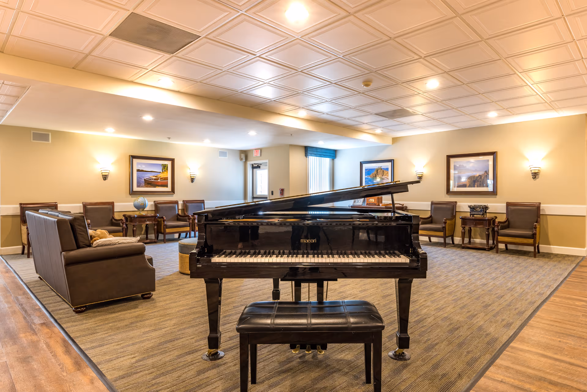 A spacious and well-lit common area featuring a black grand piano with a matching bench in the center. Surrounding the piano are several brown leather chairs and sofas arranged along the walls, with framed landscape paintings and wall sconces providing warm lighting. The room has a patterned carpet and a white coffered ceiling with recessed lights.
