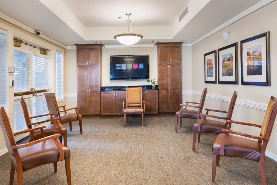 A well-lit room with six wooden chairs arranged in a semi-circle facing a wall-mounted flat screen TV. The room has beige walls adorned with three framed landscape paintings on the right side. There are two tall wooden cabinets flanking the TV, and a small vase with white flowers on the cabinet below the TV. Large windows with blinds are on the left side, and a ceiling light fixture hangs in the center of the room.
