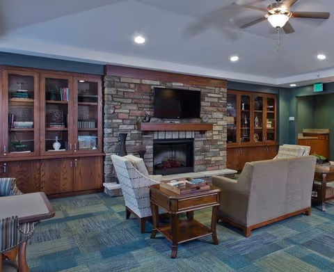 A cozy living room area featuring a stone fireplace with a mounted flat-screen TV above it. The room has built-in wooden cabinets with glass doors on either side of the fireplace, containing books and decorative items. There are comfortable upholstered chairs and a sofa arranged around a wooden coffee table. The ceiling has recessed lighting and a ceiling fan, and the floor is covered with a patterned carpet in shades of blue and green.