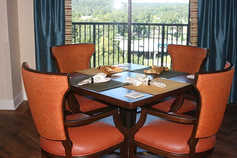 Round wooden dining table with four upholstered orange chairs set near a large window overlooking trees and a balcony.