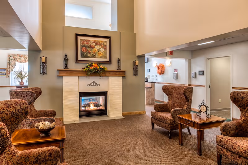 A cozy senior living facility common area with patterned armchairs arranged around wooden side tables. A fireplace with a floral painting above it is centered on the far wall, flanked by wall sconces. The room has beige walls, carpeted floor, and a high ceiling with windows near the top.