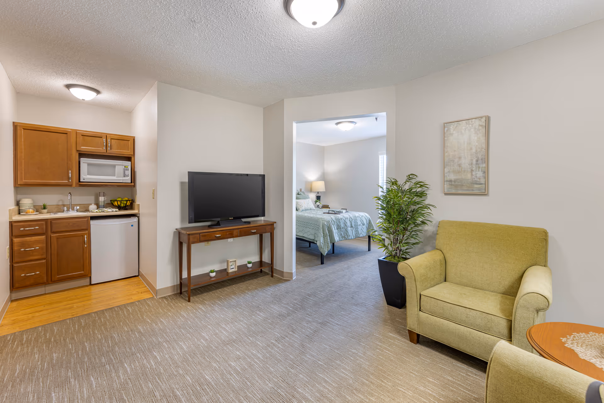 Interior view of a senior living apartment at Brookdale Findlay featuring a small kitchenette with wooden cabinets, a microwave, and a mini fridge on the left. In the center, a flat-screen TV sits on a wooden console table. To the right, there is a green armchair next to a potted plant and a framed artwork on the wall. In the background, a bedroom with a bed, nightstand, and lamp is visible through an open doorway.