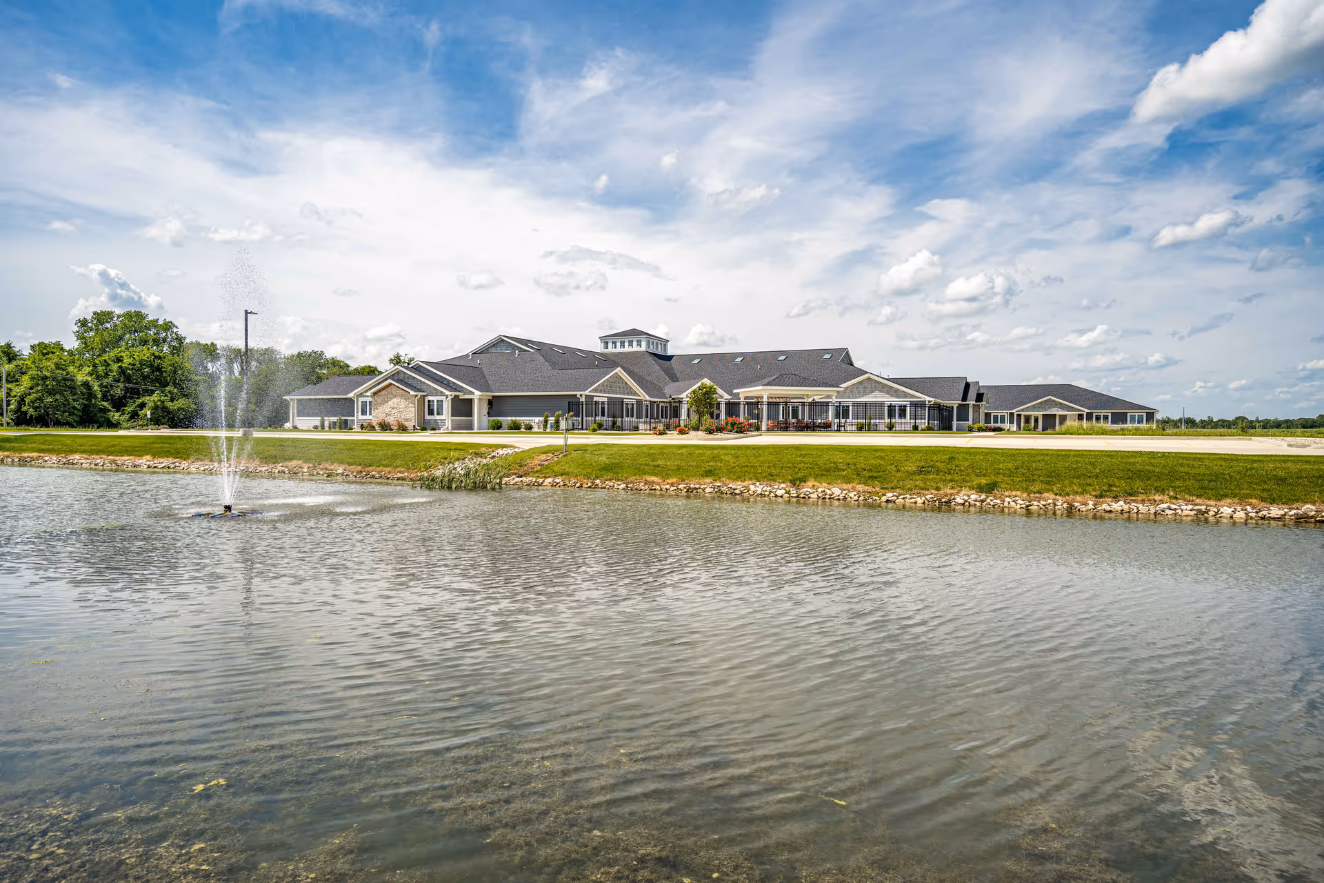 Front exterior of a long single-story assisted living building seen across a pond with a fountain under a partly cloudy sky.