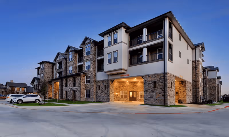 Exterior view of a multi-story senior living facility building at dusk with stone and beige siding, multiple windows, balconies, and a covered entrance. Several cars are parked in the parking lot adjacent to the building.