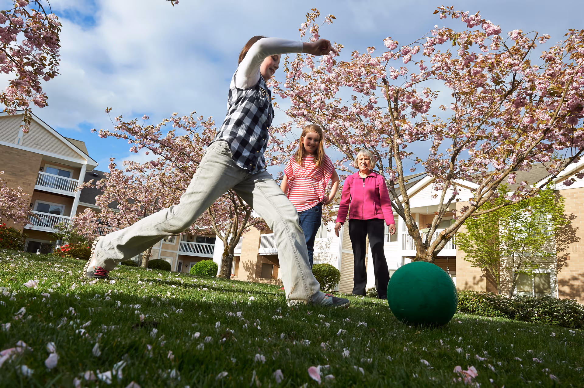 Three people playing with a large green ball on a grassy area surrounded by blooming cherry blossom trees, with residential buildings in the background under a partly cloudy sky.