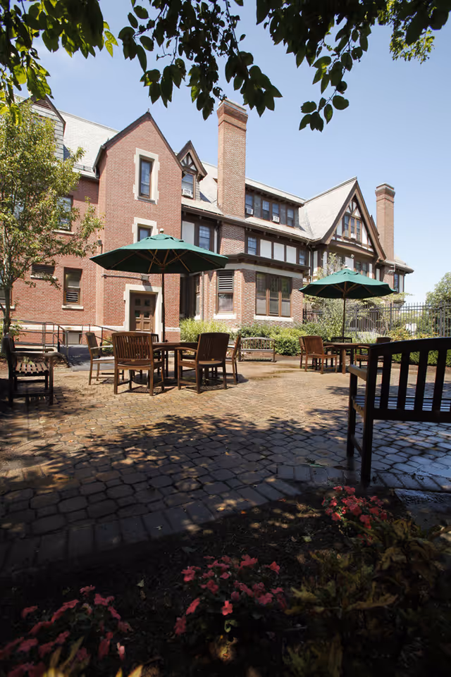 Outdoor patio area with wooden tables and chairs under green umbrellas, surrounded by a brick building with multiple windows and chimneys. There are trees and plants around the patio, and the sky is clear and blue.