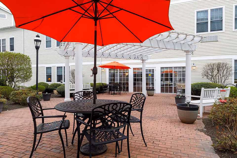 Outdoor patio area at Brookmeadow at Cobb Corner featuring a round metal table with four matching chairs under a large red umbrella. In the background, there is a white pergola, another table with an orange umbrella, white benches, potted plants, and the exterior of a light-colored building with multiple windows and glass doors.