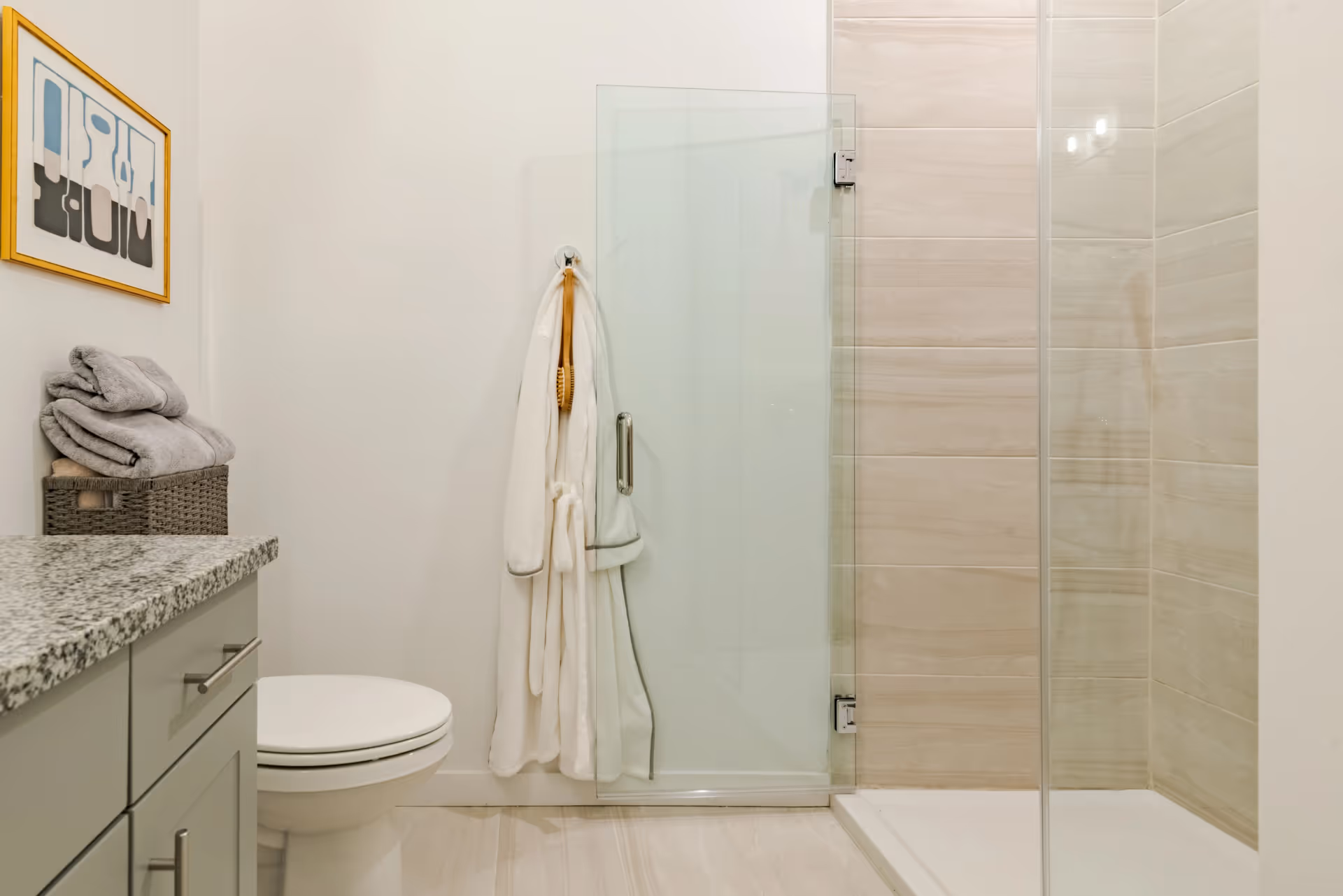 A modern bathroom featuring a toilet, a granite countertop with a basket of folded towels, a white bathrobe hanging on a hook, and a glass shower enclosure with beige tiled walls.