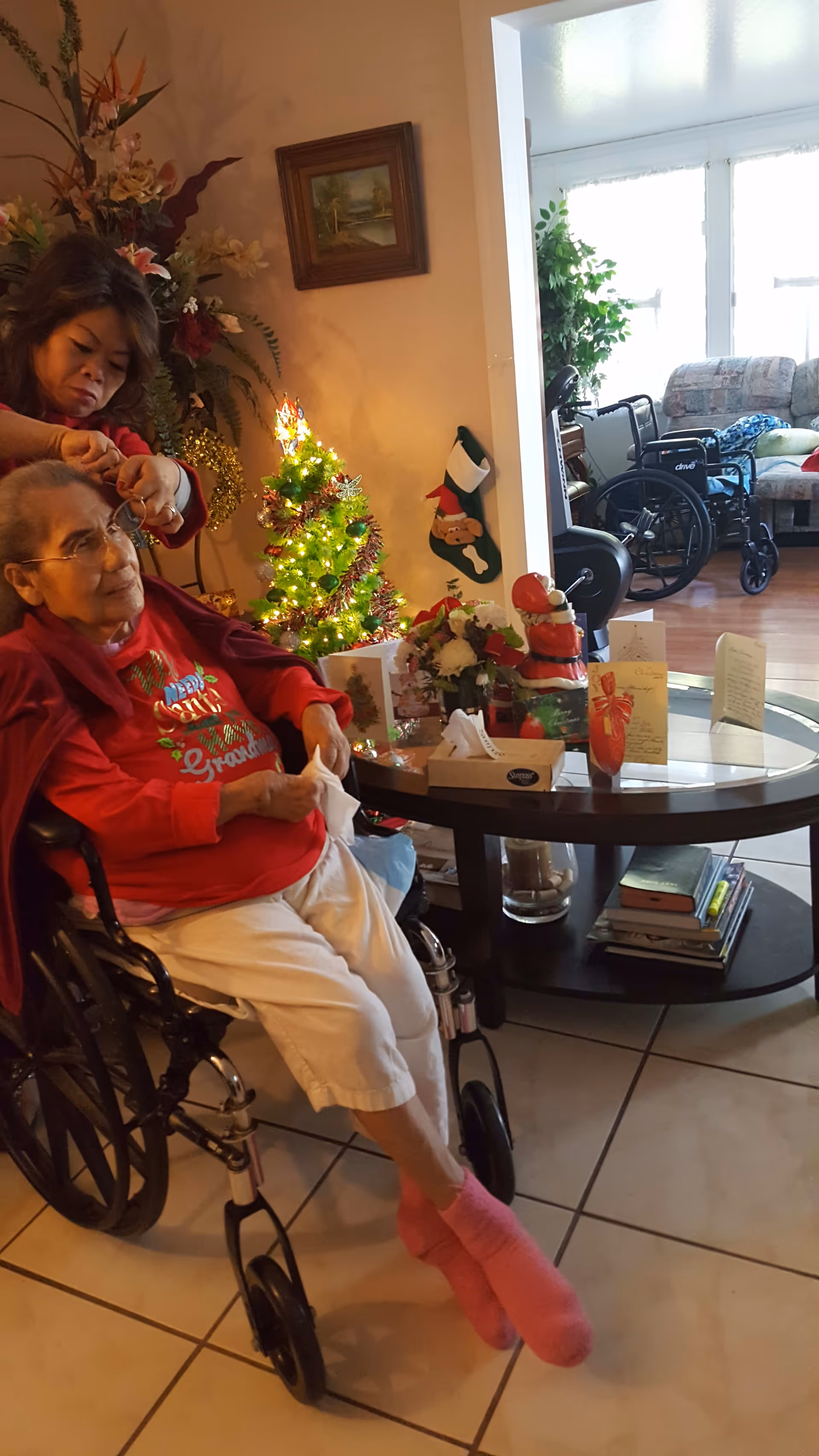 An elderly woman in a wheelchair wearing a red shirt and pink socks is having her hair styled by another woman in a cozy room decorated for Christmas with a small lit Christmas tree, holiday cards, and festive decorations on a glass coffee table. In the background, there is a living area with a couch and a wheelchair.