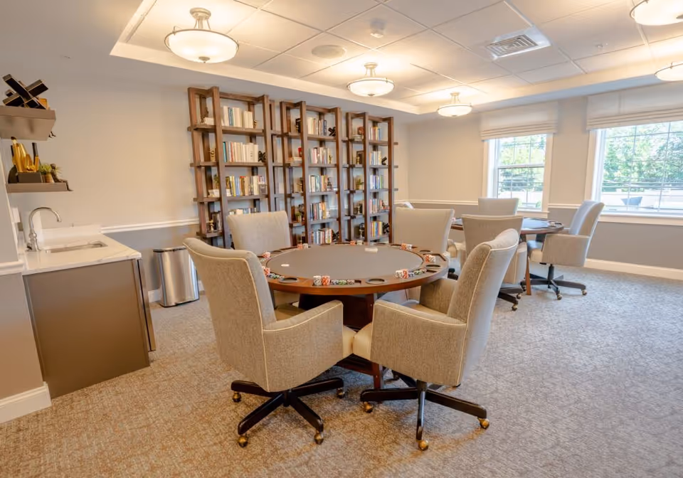 A well-lit room with two round tables surrounded by beige upholstered swivel chairs on casters. One table is set up for poker with poker chips and card holders. Behind the tables, there are tall wooden bookshelves filled with books. The room has large windows with white blinds, a small sink and countertop area on the left, and neutral-colored walls and carpet.