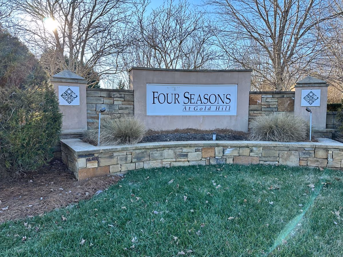 Stone entrance monument sign reading "Four Seasons At Gold Hill" with grass, landscaping, and leafless trees in the background.