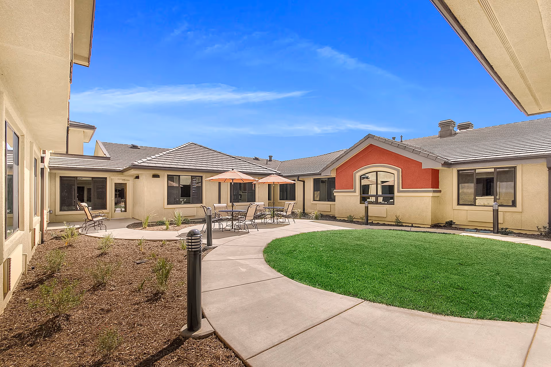 Outdoor courtyard area at The Pines, A Merrill Gardens Community, featuring a circular green lawn surrounded by a concrete walkway, patio tables with umbrellas, chairs, and beige buildings with large windows under a clear blue sky.