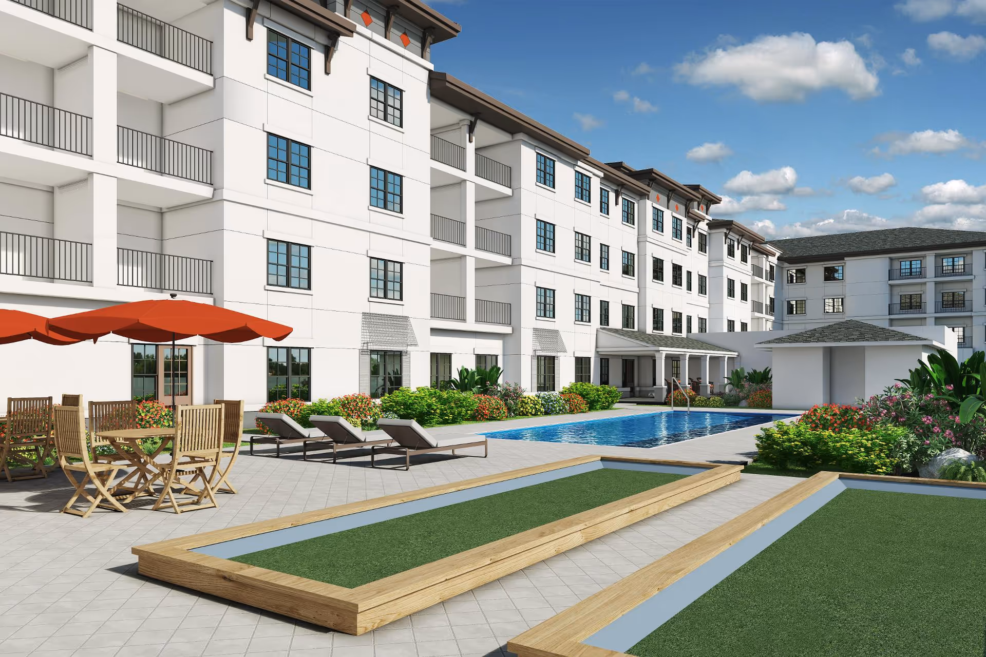 Outdoor courtyard area of a senior living facility with a swimming pool, lounge chairs, wooden tables with red umbrellas, and bocce ball courts. The building is white with multiple windows and balconies, surrounded by greenery and flowers under a partly cloudy sky.
