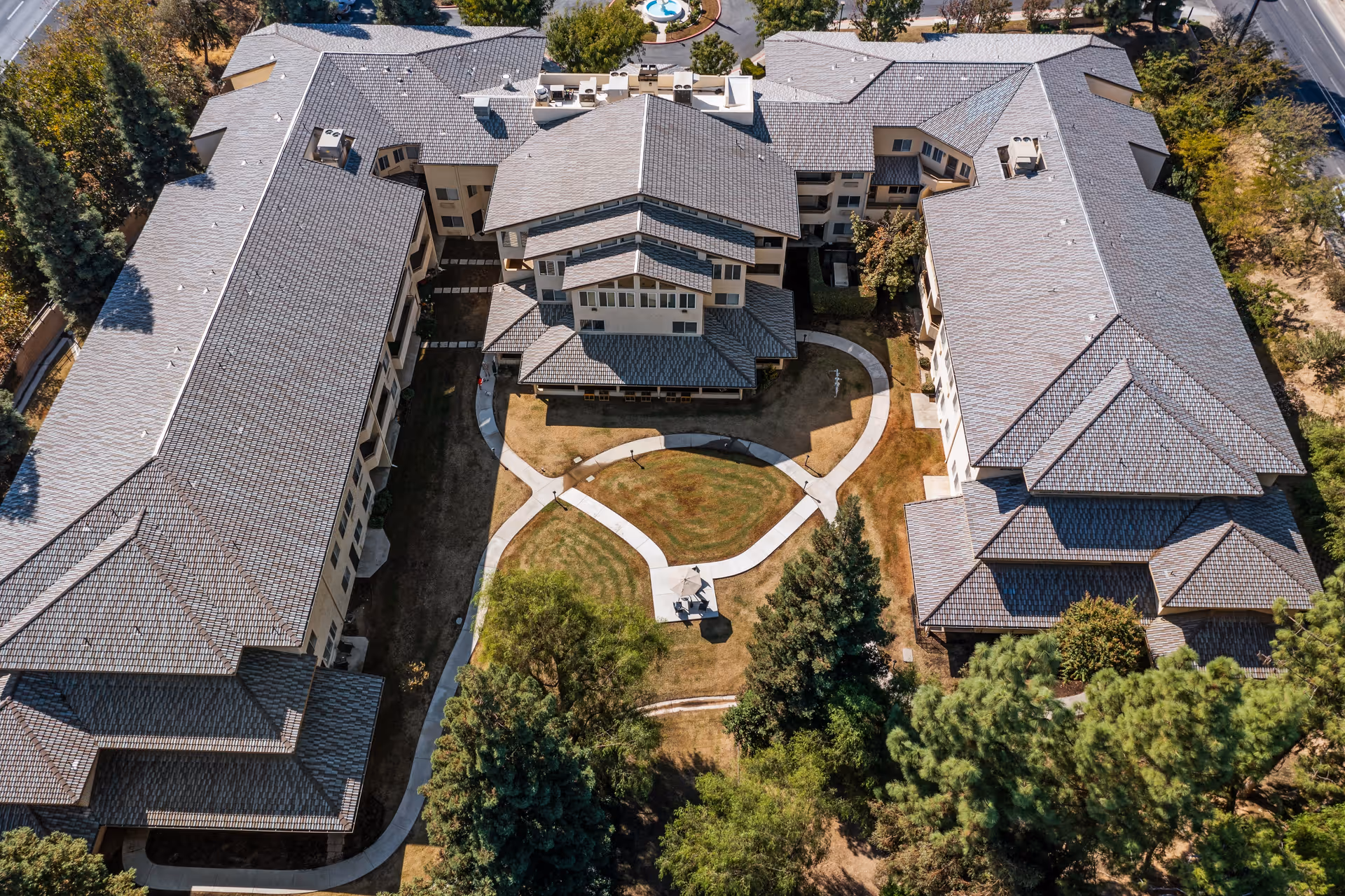 Aerial view of Columbus Estates, a senior living community, showing a large U-shaped building surrounding a central courtyard with pathways, trees, and a small seating area with an umbrella.