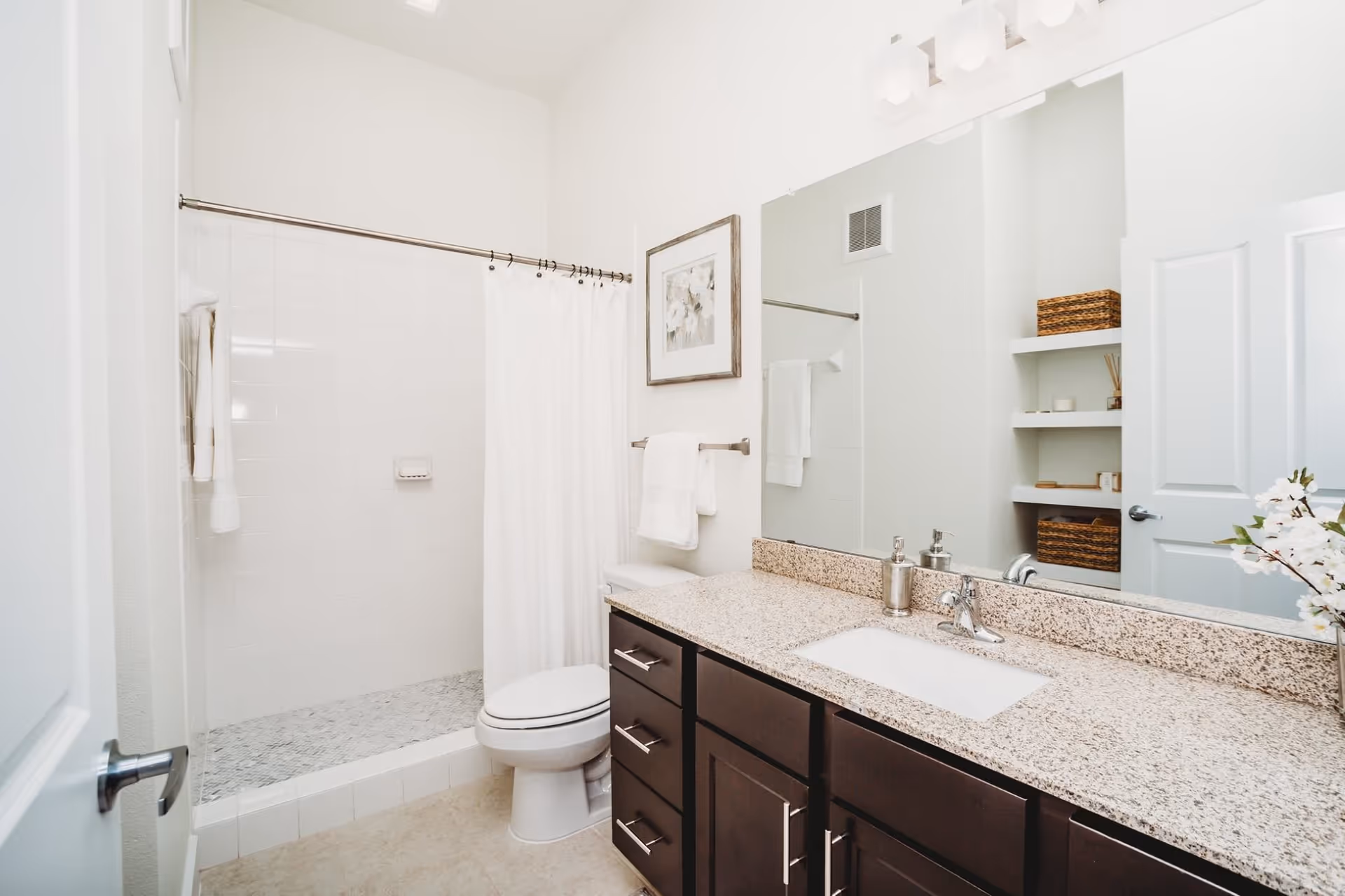Bright modern bathroom with a granite countertop vanity, large mirror, toilet, and a tiled walk-in shower with a white curtain.