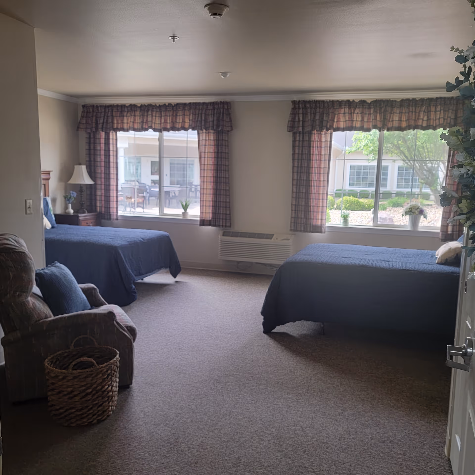A spacious bedroom with two twin beds covered in dark blue bedspreads, each positioned under large windows with plaid curtains. There is a cushioned armchair with a blue pillow and a woven basket beside it. The room has beige walls and carpeted flooring, with a heating/cooling unit beneath one window and a view of a garden and building outside.