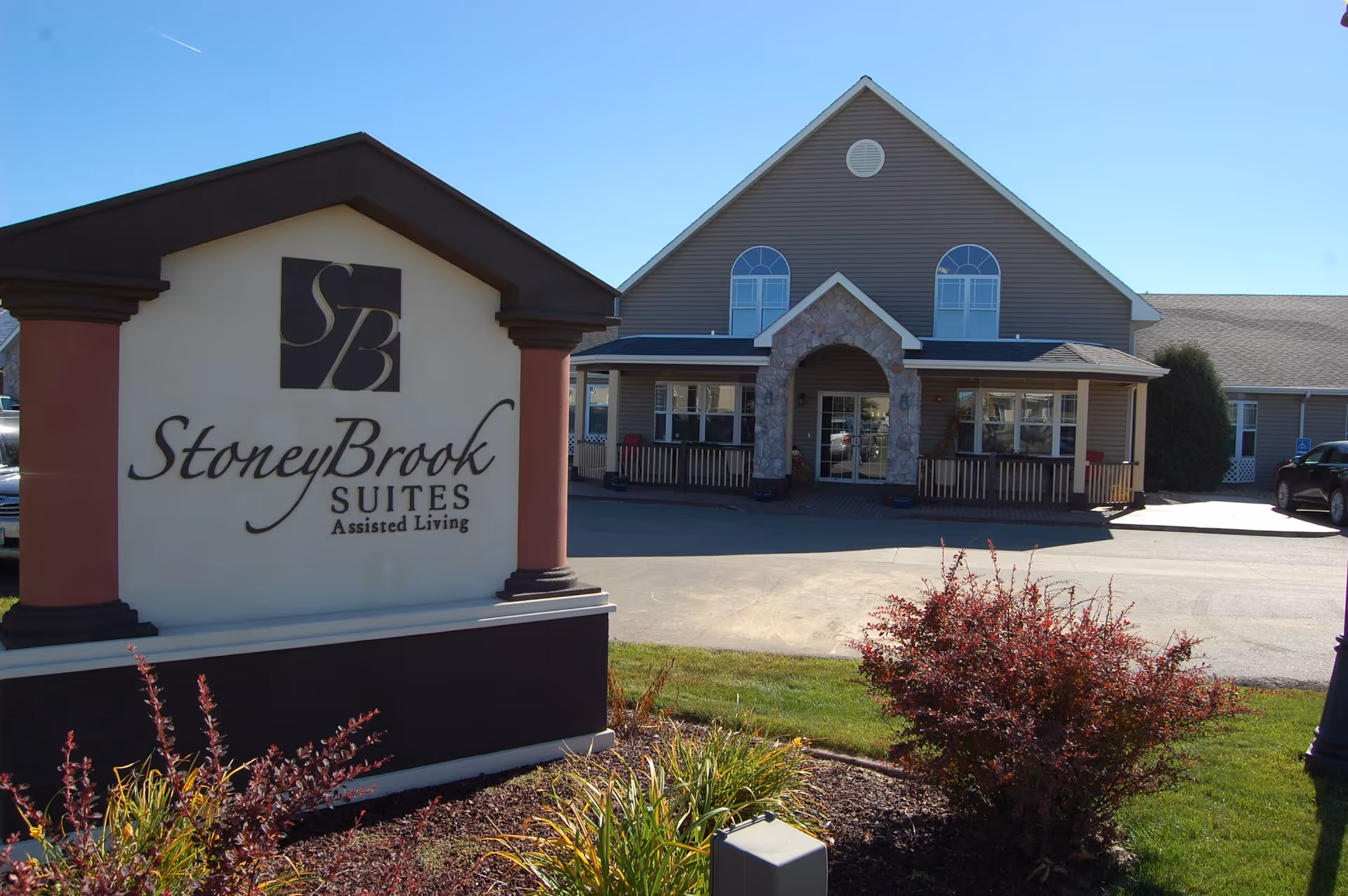 Exterior view of StoneyBrook Suites Assisted Living building with a large sign in the foreground displaying the facility's name. The building has a peaked roof, stone arch entrance, and several windows. There are shrubs and grass in the landscaped area near the sign.